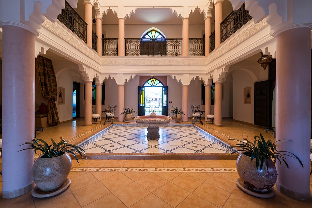 A spacious entrance hall is framed by elegant arches and columns. Natural light floods the area, highlighting decorative tiles and a central fountain surrounded by potted plants. Two large doors lead outside, offering a view of the lush surroundings.