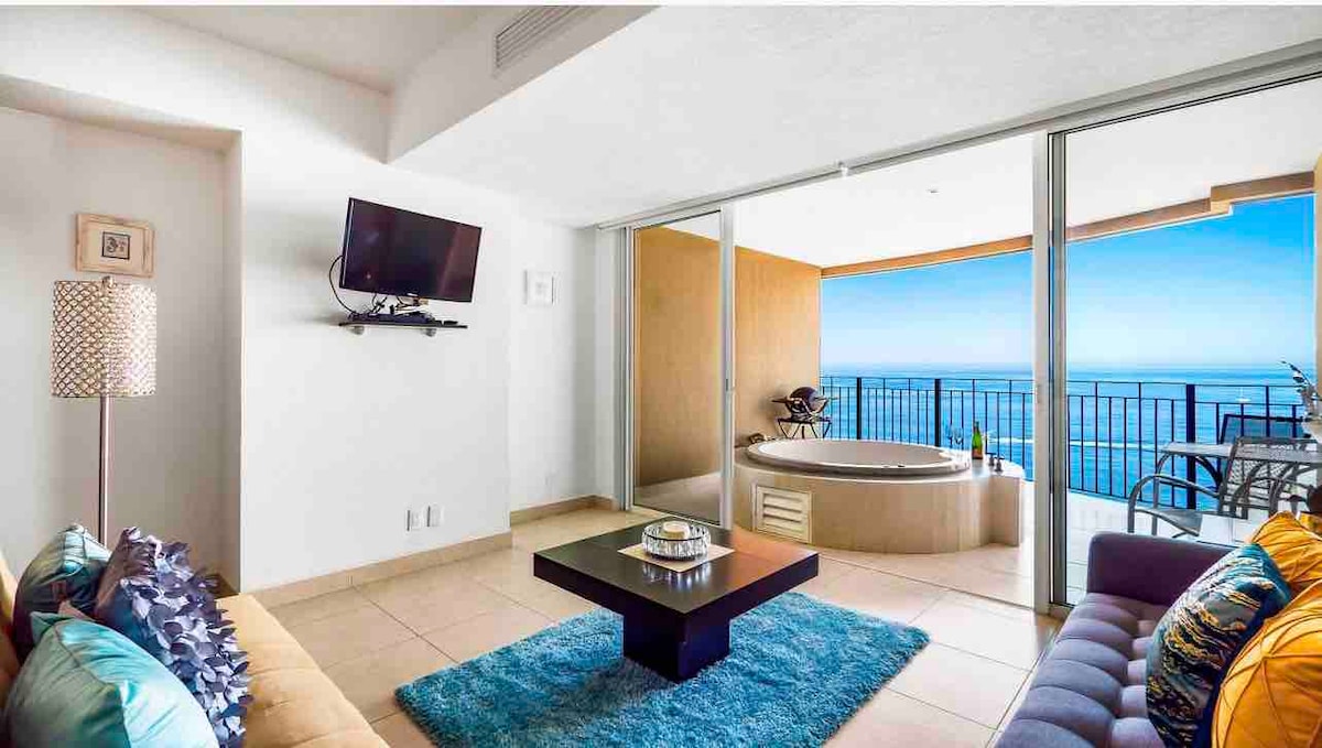 A welcoming living area features a contemporary design with a dark coffee table at the center. A private balcony jacuzzi is visible through expansive glass doors, offering unobstructed views of the ocean. Colorful cushions adorn the seating, providing comfort and inviting relaxation.