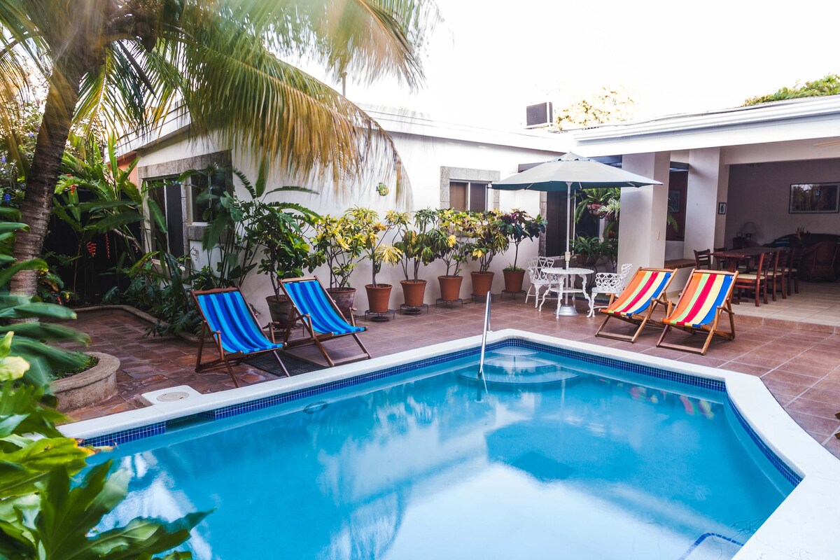 A refreshing pool is surrounded by a patio area featuring striped lounge chairs. Lush greenery and potted plants create a serene atmosphere. An umbrella provides shade over a seating area, while nearby windows indicate the presence of indoor living spaces.