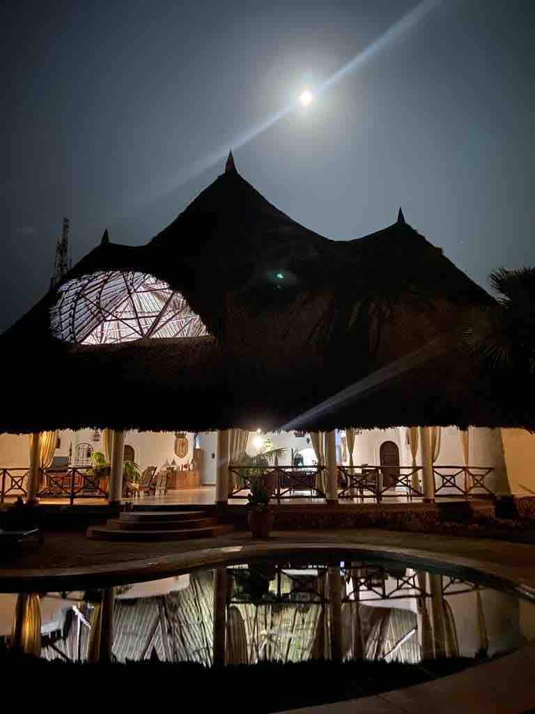 A beautifully crafted coastal villa is illuminated under a night sky, showcasing a distinctive palm leaf roof. Reflections of the structure are visible in the tranquil salt-water pool, creating a serene atmosphere. Moonlight enhances the scene, highlighting the architectural details and surrounding tropical greenery.