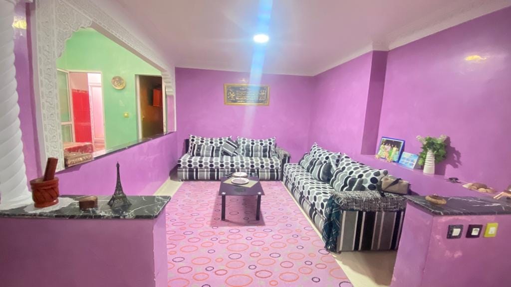 A colorful living room features two striped sofas, complemented by a small table with decorative items. The walls are painted in vibrant purple, and a patterned rug covers the floor. Soft lighting illuminates the space, enhancing its welcoming atmosphere.