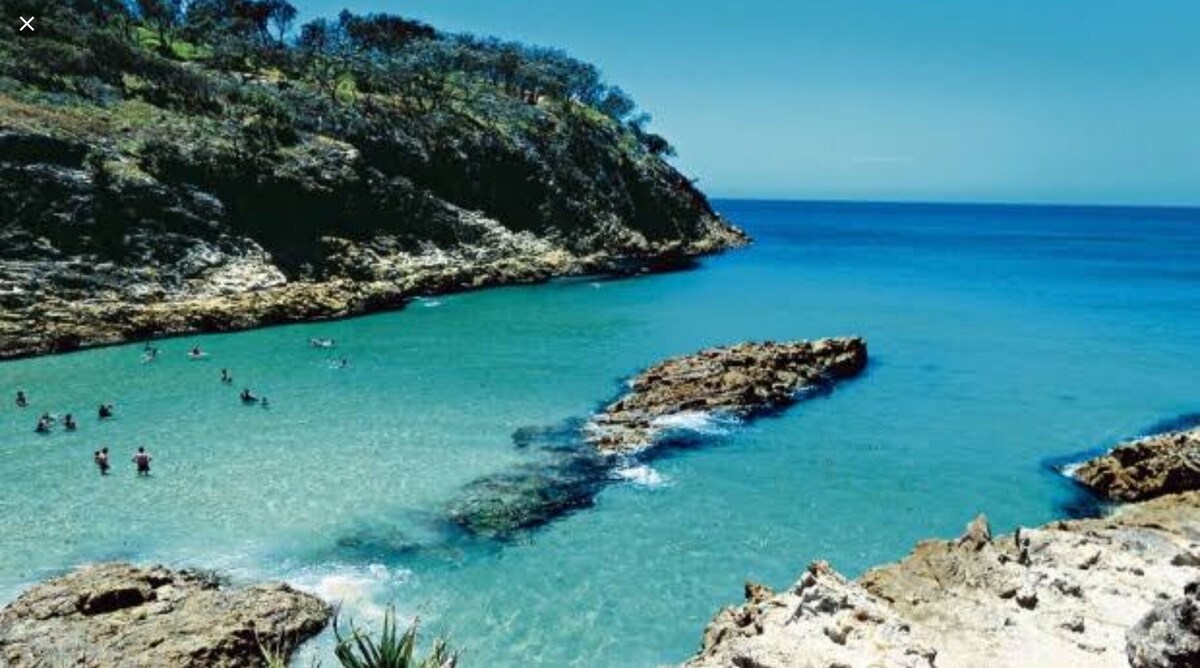 A serene coastal scene showcases a calm bay surrounded by rocky shorelines and lush greenery. The clear blue water invites swimmers, while gentle waves lap against the rocks. The horizon reveals a seamless blend of ocean and sky, creating a tranquil atmosphere.