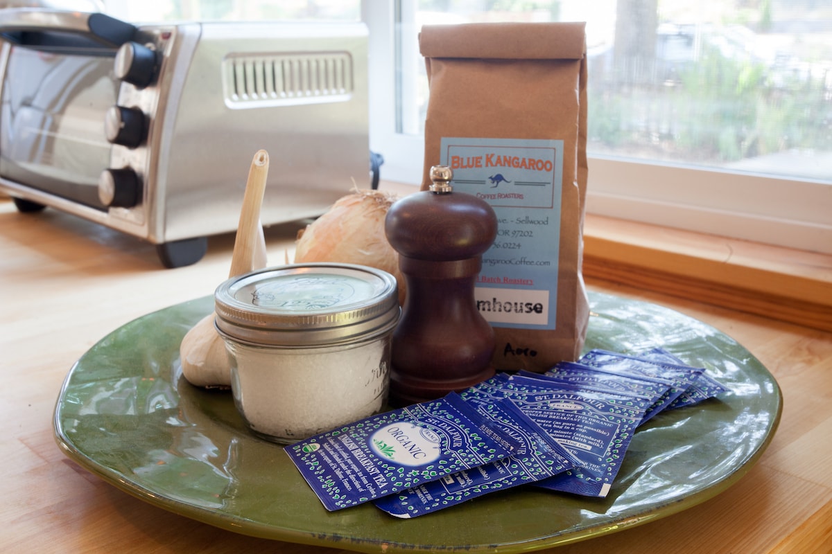 A green ceramic platter displays various cooking essentials, including a jar of salt, a silver pepper mill, and a bag of coffee. Several small packets are arranged beside the other items, complementing the kitchen's natural light streaming in through a nearby window.