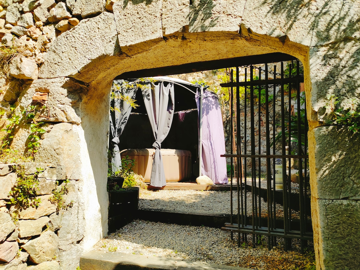 An entrance framed by rustic stonework leads to a private outdoor area. A jacuzzi is partially visible, sheltered by elegant gray curtains. Pebbled ground surrounds the space, creating a serene environment. Lush greenery adds a natural touch to the setting.