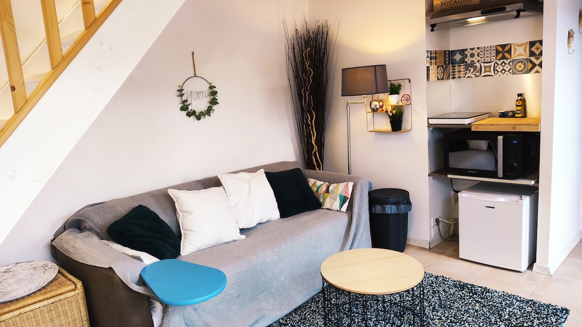 A cozy living area features a spacious couch adorned with cushions and a soft throw. A small coffee table sits on a textured rug, and a kitchenette is visible in the background with a microwave and fridge. A decorative wall hanging adds a personal touch.