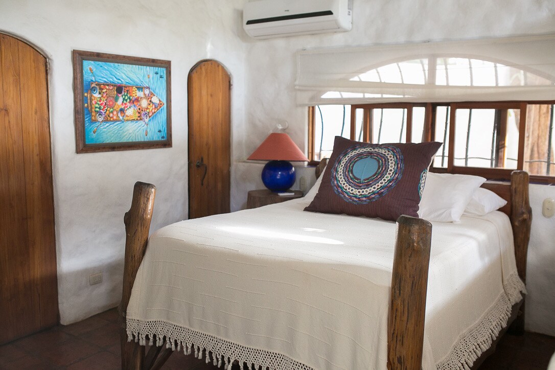 A cozy bedroom features a spacious bed with a white coverlet and decorative pillows. Natural wood elements are used for the bed frame and doors. Light filters through the window, enhancing the soft textures and an art piece depicting a colorful boat is displayed on the wall.