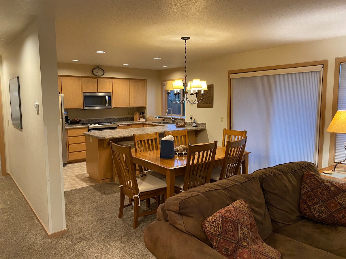 An open dining area is presented, featuring a wooden table that comfortably seats six. Nearby, a well-equipped kitchen is visible, with light wood cabinetry and a countertop. Soft lighting is provided by a pendant fixture and floor lamp, creating a cozy ambiance within the space.