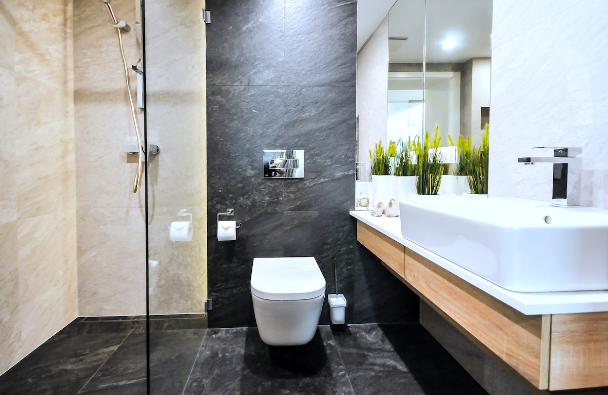 The modern bathroom features sleek stone walls and a minimalist design. A walk-in glass shower is visible alongside a contemporary toilet and wooden vanity. A large mirror reflects the space, while decorative plants add a touch of greenery to the room.