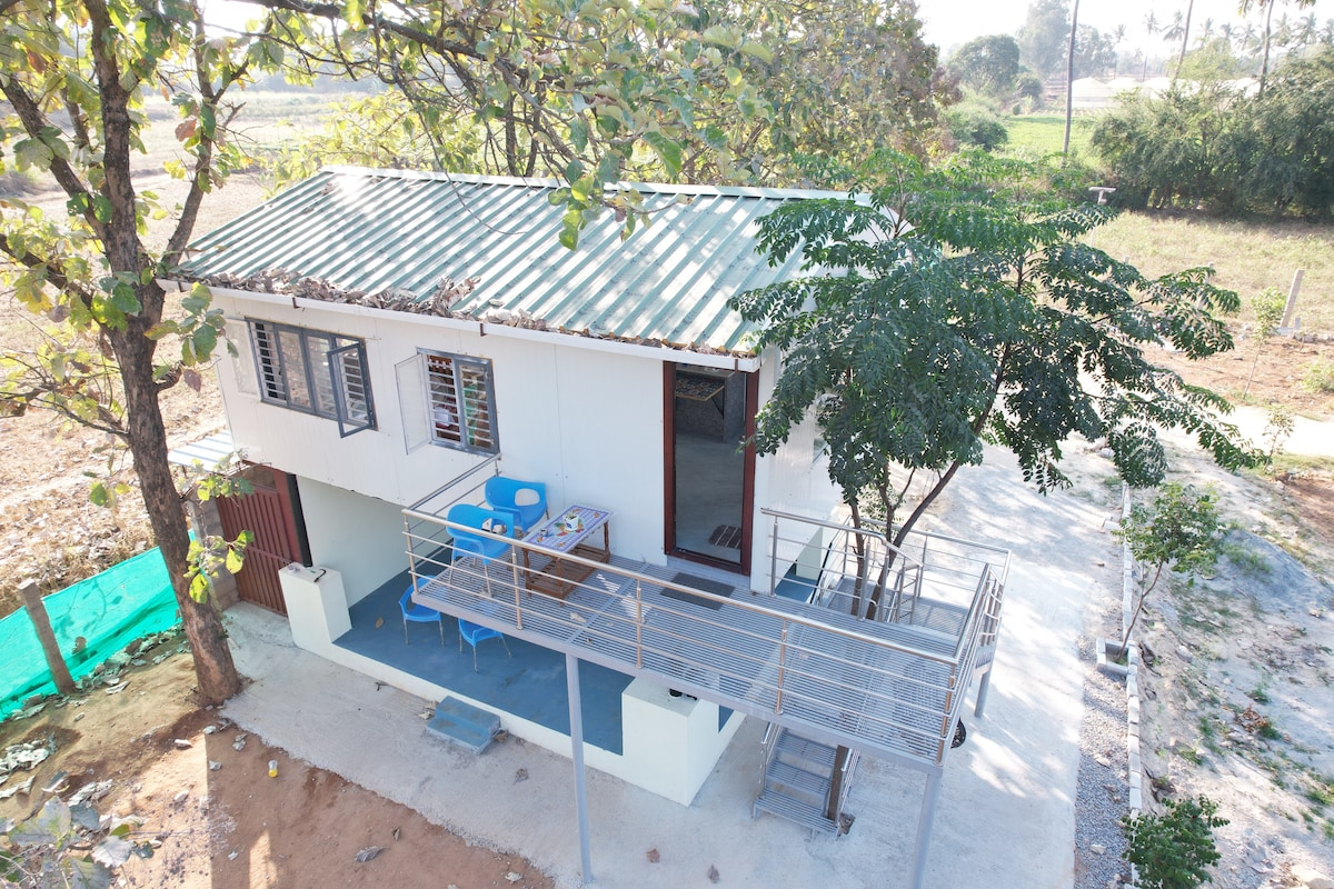 A quaint farmhouse is featured, surrounded by greenery. A spacious balcony with seating is visible, offering views of the landscape. The structure is elevated, showcasing a slanted roof and multiple windows that invite natural light into the interior.