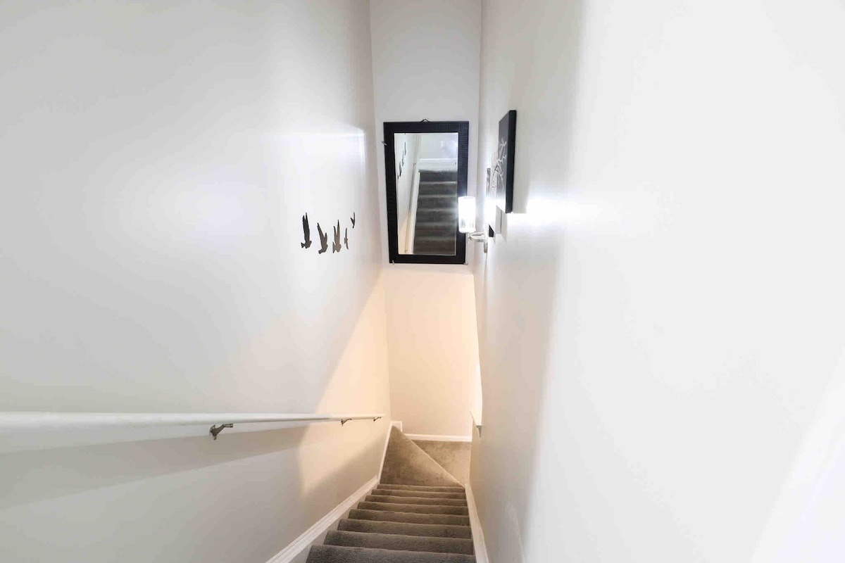 A light-filled staircase is highlighted by soft gray walls and plush carpet. Wall decor featuring silhouettes is present beside the staircase. A mirror adds depth to the space, while warm lighting illuminates the area, guiding the way to the upper level.