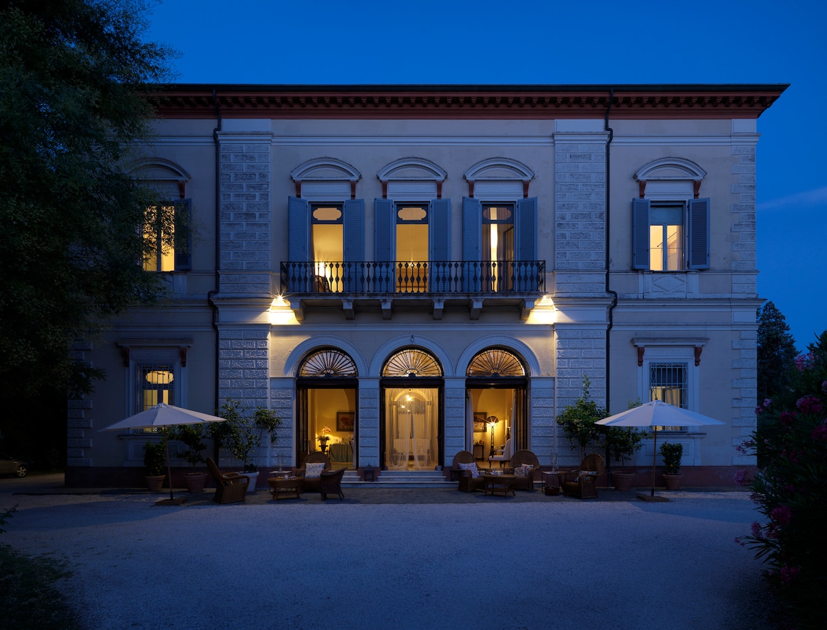 The historic villa stands elegantly at dusk, featuring arched windows adorned with decorative shutters. Warm light spills from the interior, enhancing the serene ambiance. Outdoor seating is provided under large umbrellas, inviting relaxation in the charming courtyard surrounded by lush greenery.