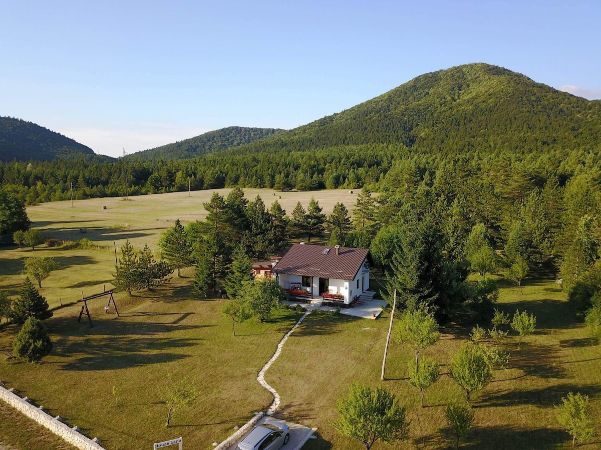 The holiday home is set within a spacious garden, surrounded by lush greenery and trees. Rolling hills are visible in the background, enhancing the serene countryside atmosphere. A swing set is located nearby, indicating a family-friendly environment.