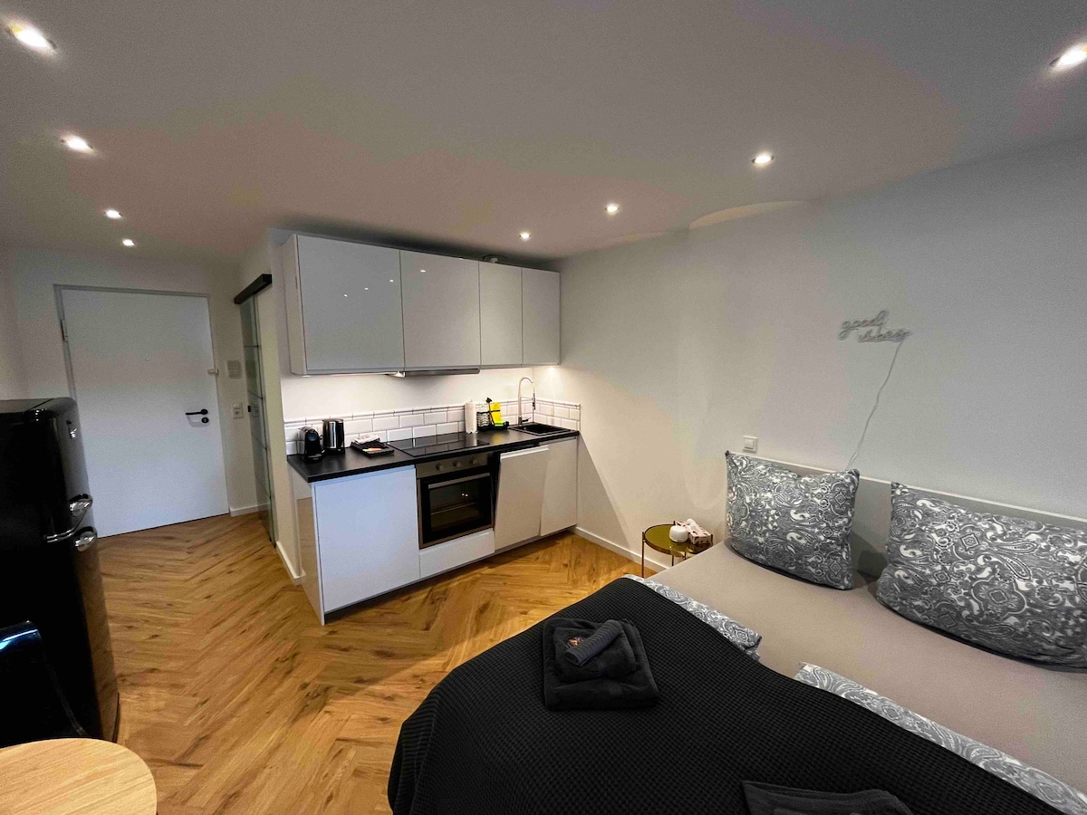 A modern open-plan space features a well-equipped kitchen with sleek cabinets and a counter. A cozy sleeping area is complemented by textured pillows on the bed. Warm lighting creates a comfortable ambiance, while wooden flooring adds a natural touch.