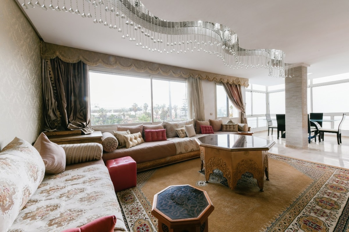 A spacious living area features a traditional Moroccan-style sofa adorned with decorative cushions. A central coffee table with intricate design is positioned on an ornate rug. Large windows provide natural light, showcasing views of the surroundings, while a unique chandelier adds elegance to the room.