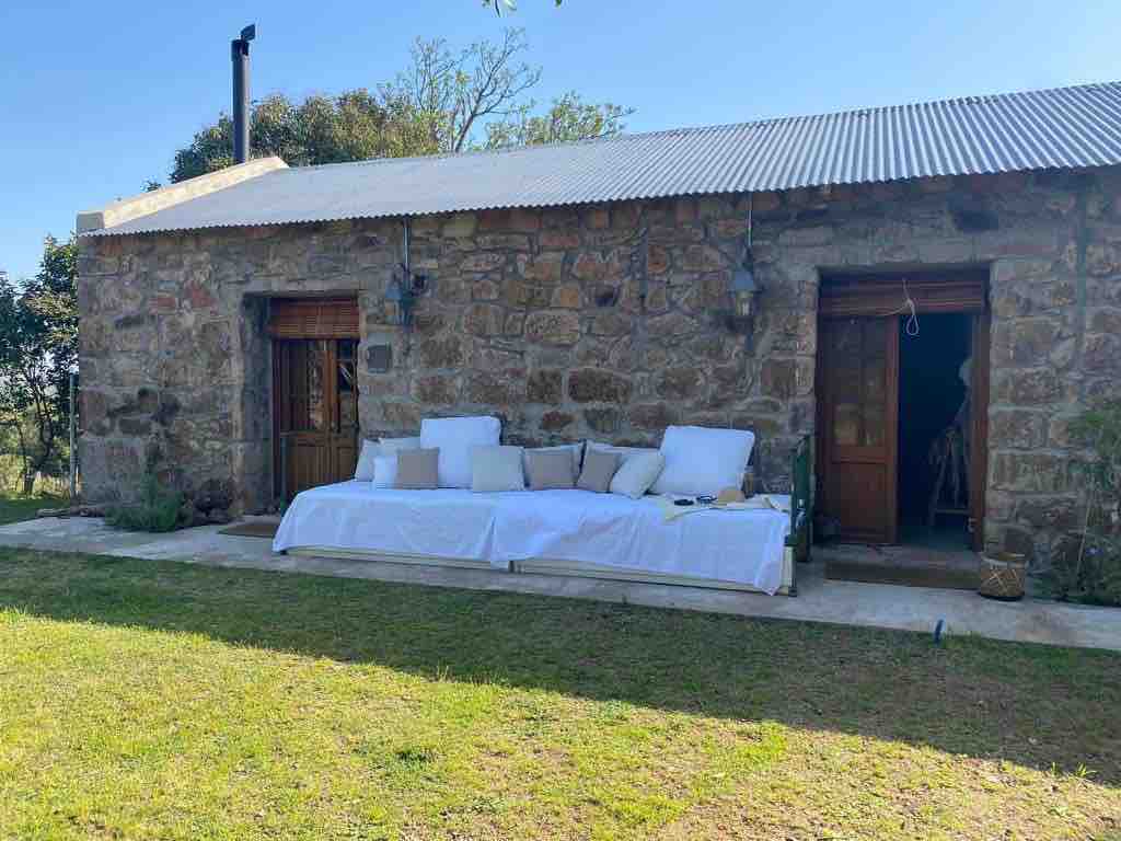 A stone exterior is featured on this rustic cabin, complemented by wooden doors. A long seating area with white cushions is positioned on a concrete patio, surrounded by grassy space and native plants. The rooftop is metallic and the landscape includes trees.