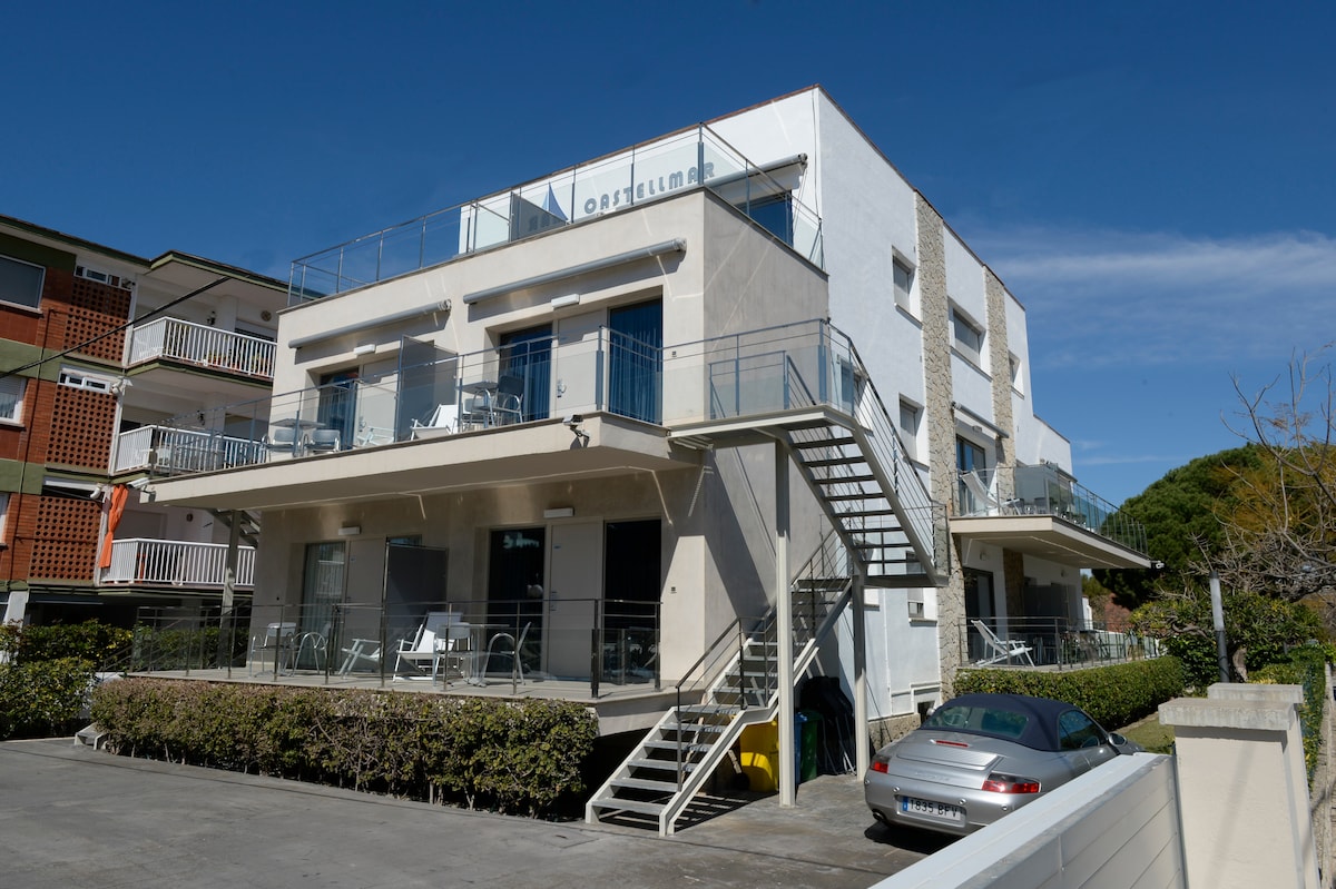 A modern three-story building is showcased, featuring multiple balconies and large glass windows. The ground level includes outdoor seating areas. A stairway on the side provides access to upper floors, while a parked car is visible in the driveway adjacent to well-maintained greenery.