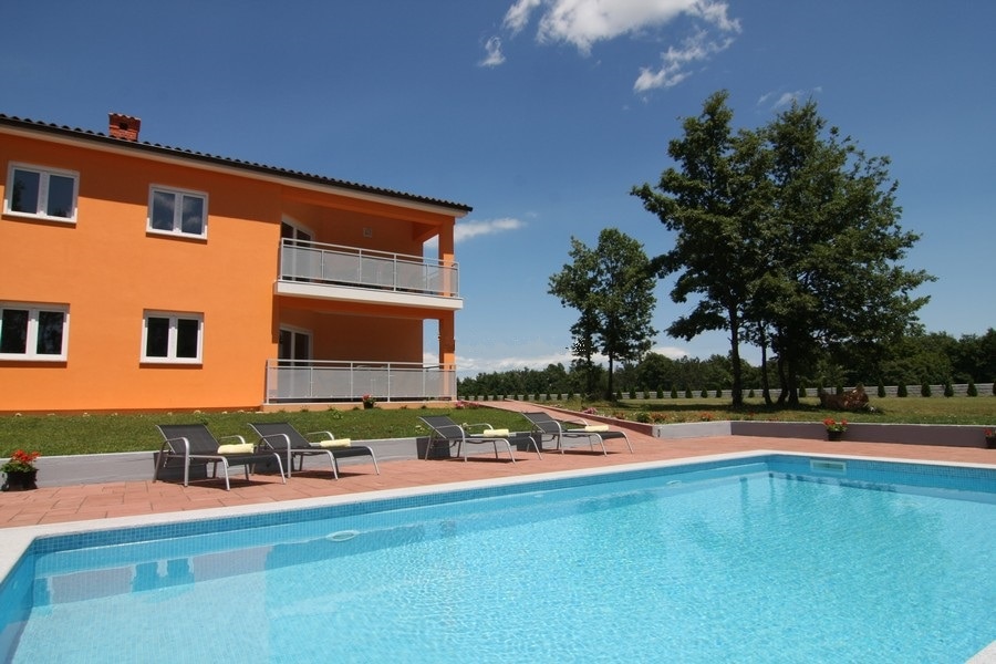 The exterior of a vibrant orange building is seen next to a clear swimming pool. Comfortable lounge chairs are lined around the pool area, complemented by potted plants. Lush greenery and a tranquil environment are present in the background, enhancing the peaceful setting.