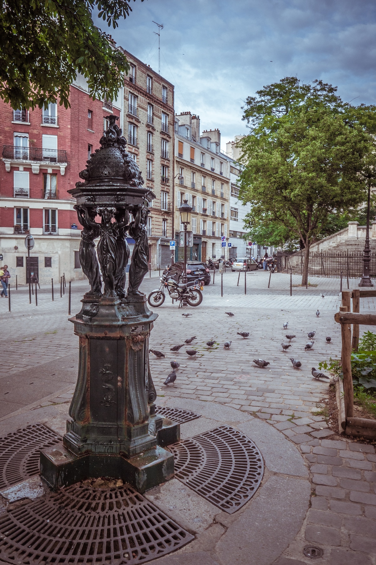 A vintage public drinking fountain is situated on the corner of a tree-lined square. Surrounding structures exhibit classic Parisian architecture, with details of wrought iron and brick. Pigeons gather around the cobblestone area, while a motorcycle is parked nearby, adding a touch of urban life.