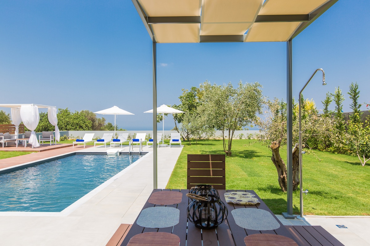 An outdoor dining area is set beneath a shaded pergola, featuring a wooden table adorned with placemats. A refreshing swimming pool stretches into the background, bordered by sun loungers and shaded umbrellas. Lush greenery and olive trees surround the space, enhancing the serene atmosphere.
