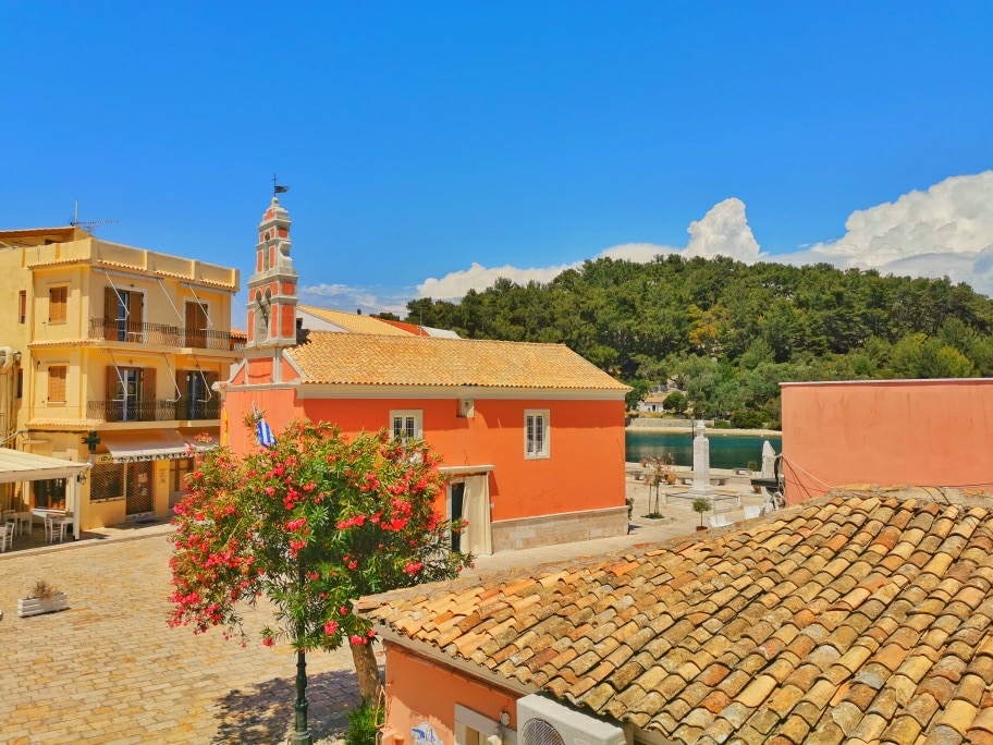 A vibrant view of the island square captures colorful buildings, including a pink structure with a clock tower. Lush greenery is visible in the background, while a blue sky with scattered clouds adds to the scenery. This image highlights the charm of the surroundings.