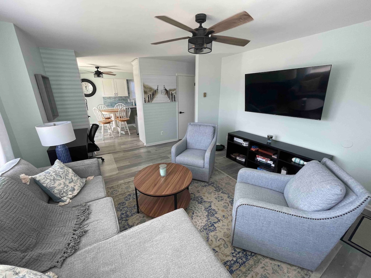 A comfortable living area is equipped with a sectional sofa and two armchairs, arranged around a round coffee table. A mounted television is positioned on the wall, with a bookshelf beneath it. Light colors and a ceiling fan contribute to the space's relaxed ambiance.