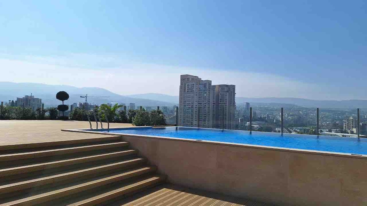 An infinity pool is set against a backdrop of urban buildings and distant mountains. Steps lead down to the pool area, which is surrounded by greenery. The clear sky enhances the serene atmosphere of the outdoor space.