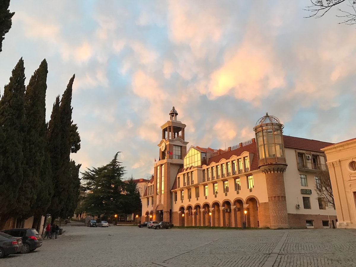 A historic building is featured in the image, characterized by a clock tower and large, arched windows. The surrounding area includes cobblestone pathways and tall trees, with soft pastel colors of the clouds in the sunset sky. Vehicles are parked nearby.