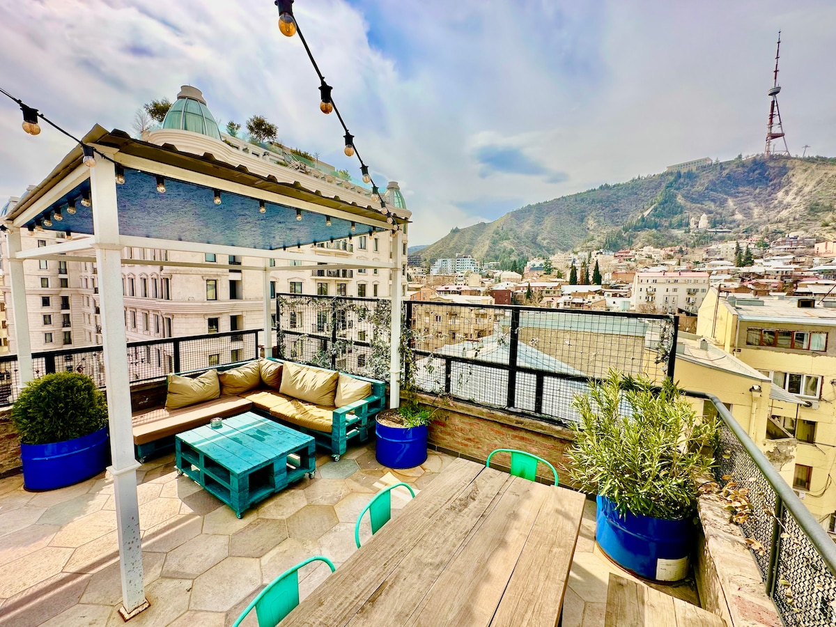 A shared terrace space features comfortable seating arrangements and a dining table. Potted plants add a touch of greenery, while views of the surrounding city and the distant mountain are visible. Outdoor string lights enhance the setting, creating a welcoming atmosphere for relaxation.