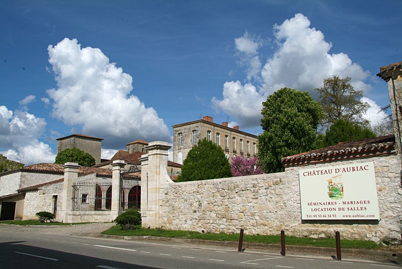 The image showcases the Château d'Aubiac, a historic stone building set against a backdrop of fluffy white clouds. A large gated entrance is framed by mature trees, with a sign indicating event and venue services. The architecture reflects a traditional style typical of the region.