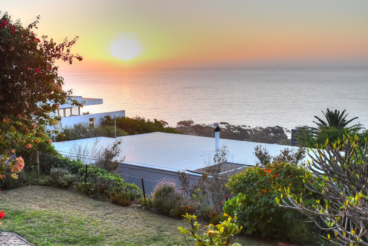 A scenic view captures the ocean at sunset, displaying vibrant shades of orange and pink across the sky. Lush greenery and colorful flowers frame the foreground, with a hint of residential structures nestled along the coastline in the background.