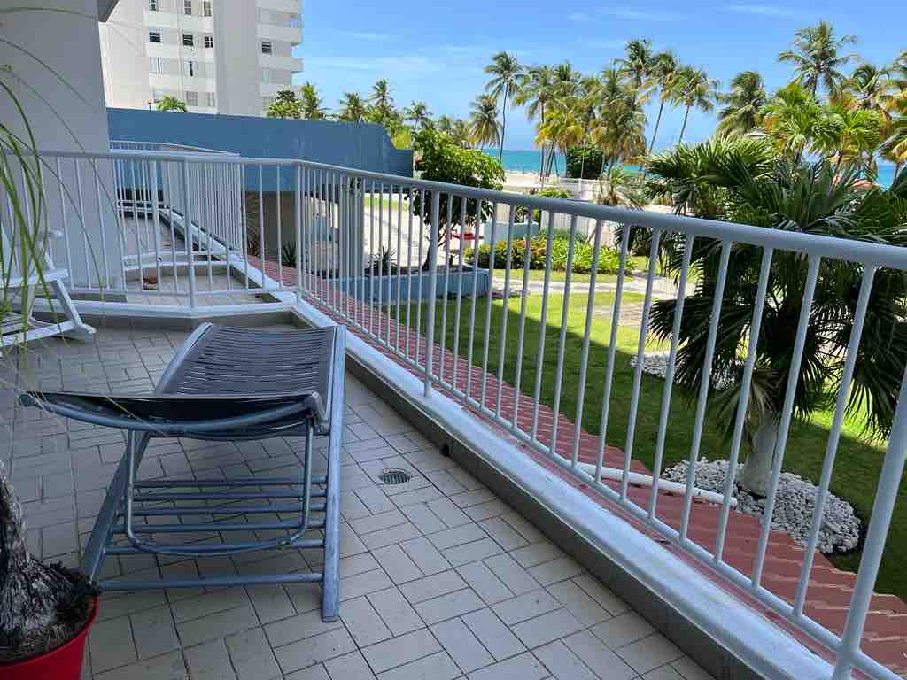 A spacious balcony features a lounge chair positioned for relaxation. Lush palm trees are visible beyond the railing, with a clear view of the beachfront and ocean in the distance. The flooring is tiled, complementing the contemporary layout.