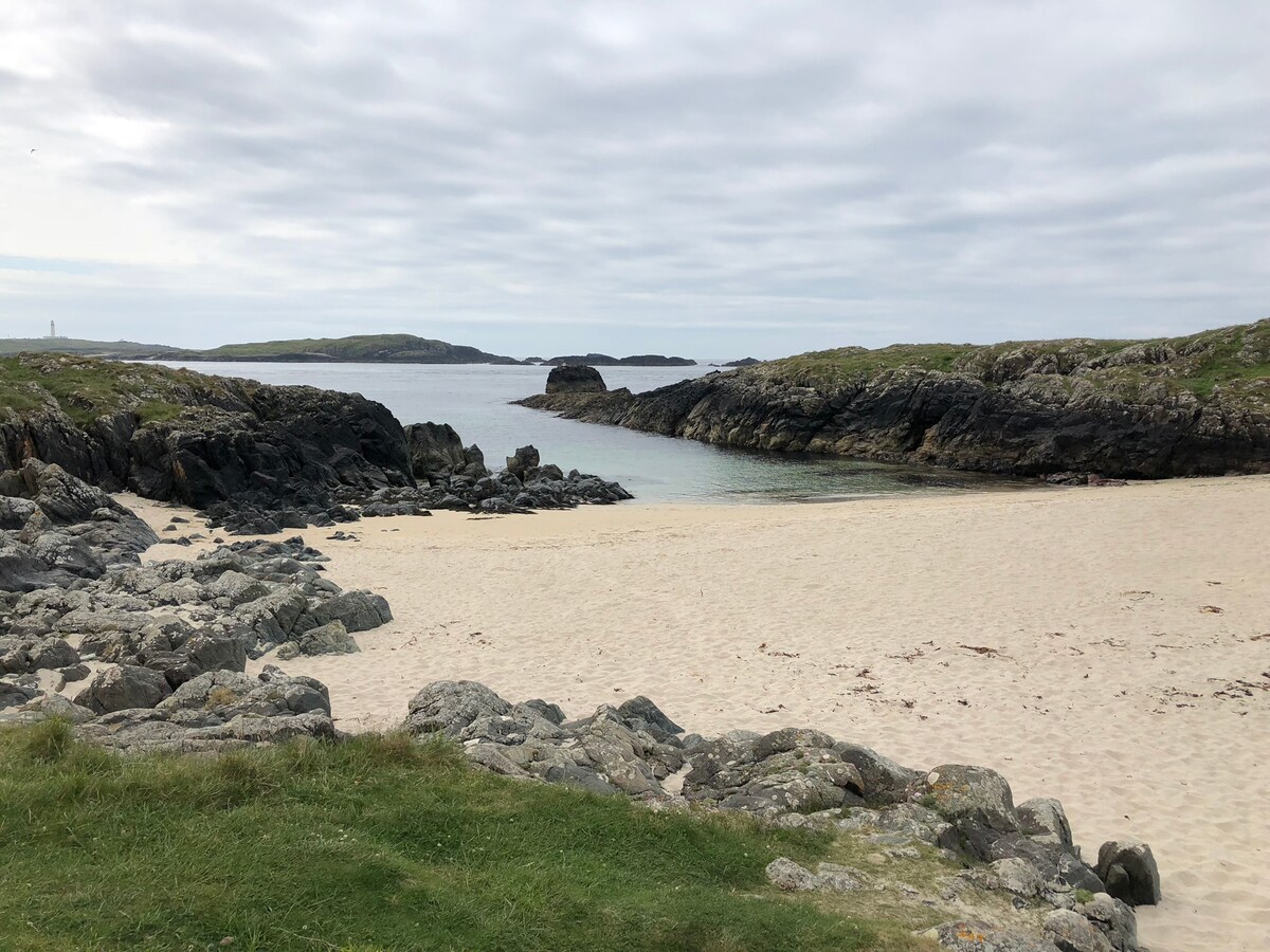 A sandy beach is framed by rocky outcrops, providing a serene coastal setting. Gentle waves lap at the shore, while grassy areas lead to the sand. The landscape is dotted with islands in the distance, under a cloudy sky.