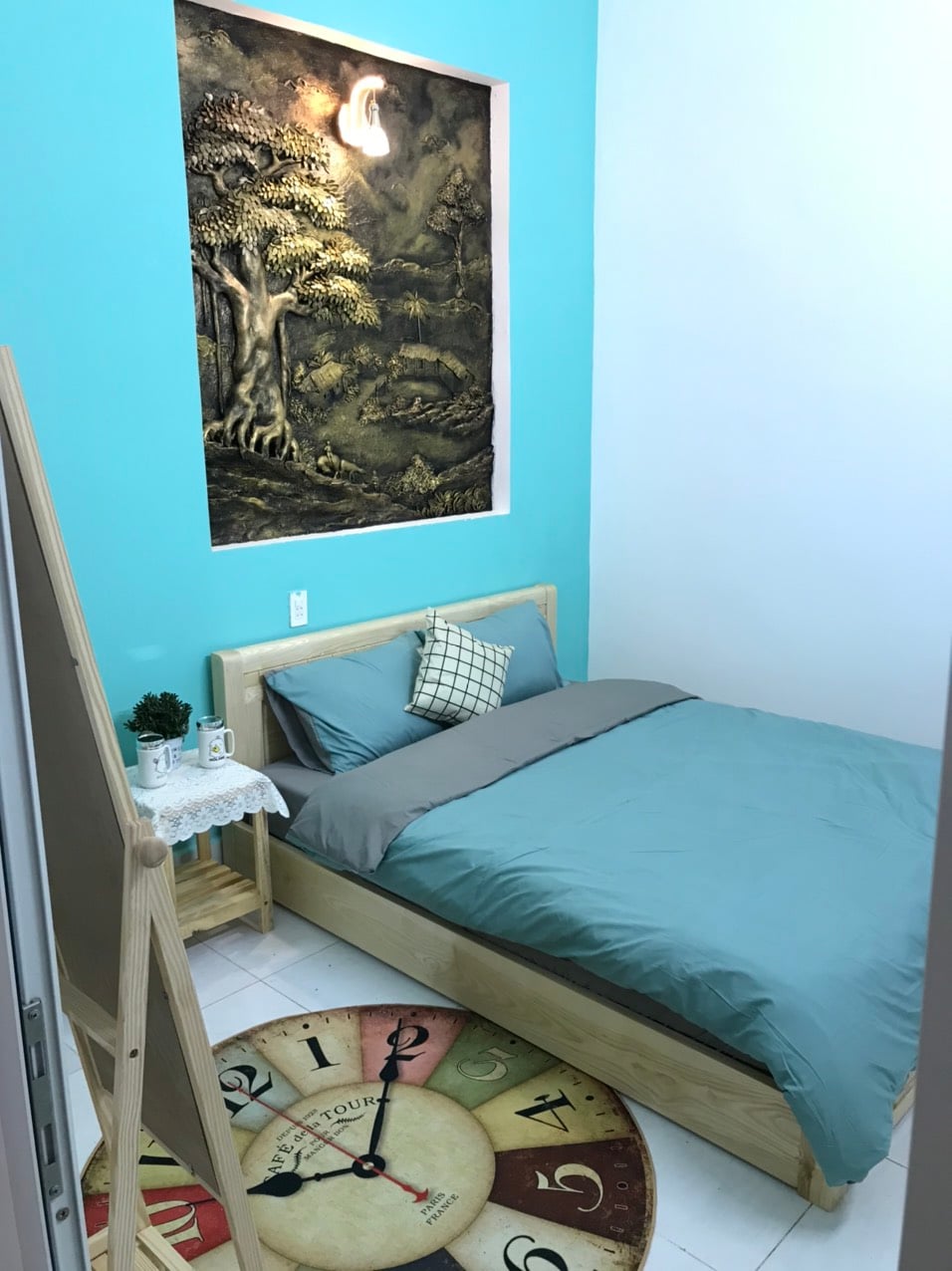A sleeping area features a wooden bed with a light blue comforter and a patterned pillow. An art piece depicting a tree is mounted on an aqua wall. A round rug with a clock design is placed on the floor, and a small plant is positioned on a side table.