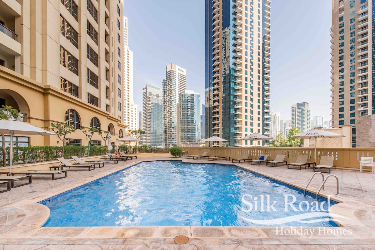 An outdoor swimming pool is surrounded by sun loungers and umbrellas, set against a backdrop of tall modern buildings. The pool's clear blue water reflects the surrounding architecture, and landscaped greenery is present along the edges.