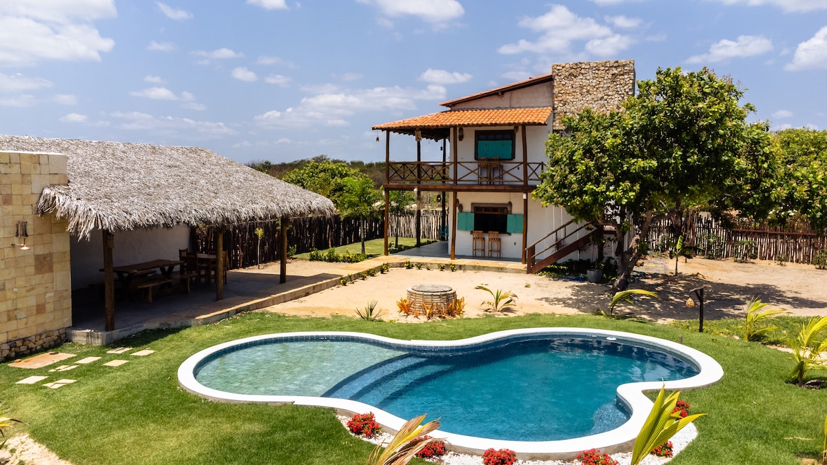 A two-story house is set amidst a spacious outdoor area, featuring a uniquely shaped pool surrounded by lush grass. Nearby, a thatched-roof structure offers shade, while seating areas are positioned to enjoy the serene surroundings.