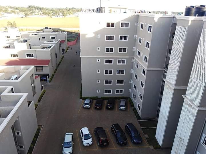The image depicts a view of a modern apartment complex, showcasing well-maintained buildings with multiple windows. A spacious parking area is visible, accommodating several cars. The surroundings appear green and open, indicating a serene atmosphere, complemented by the layout of the nearby structures.