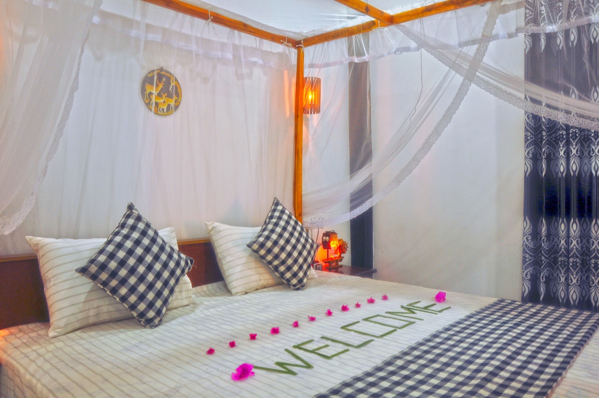 A spacious bed is framed by a wooden canopy, with decorative mosquito netting gently draped above. Checkered pillows are placed at the head of the bed, and the word 'WELCOME' is spelled out with flower petals on the linens. A wall clock and ambient lighting enhance the calming atmosphere.