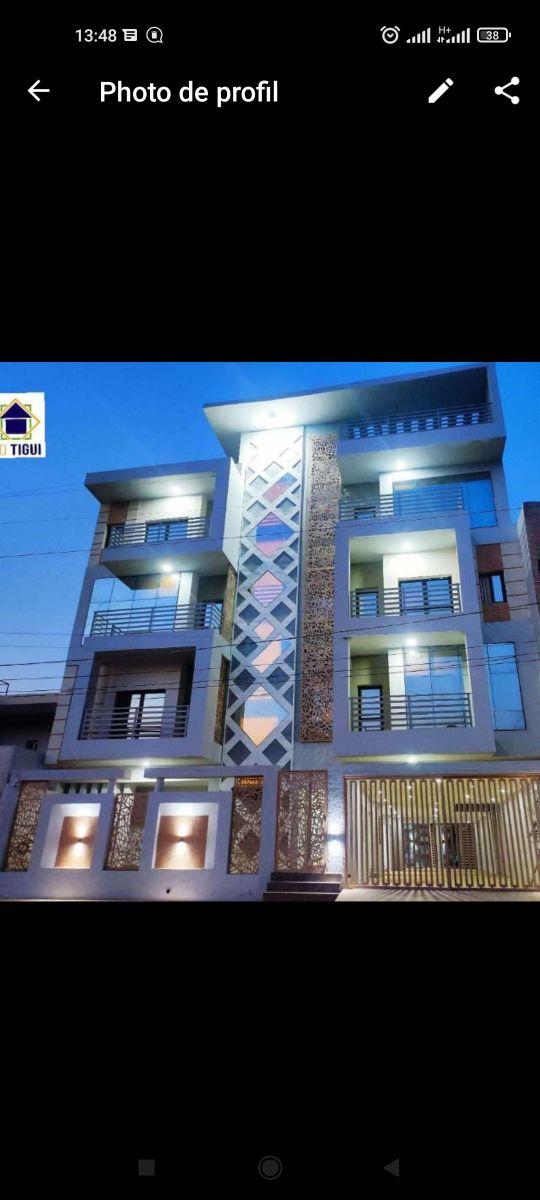 A modern three-story building showcases a geometric façade with decorative elements, illuminated at night. Large balconies are positioned on the upper levels, complemented by spacious windows. Ground-level lighting creates a welcoming entrance, while a gated area offers security and access.