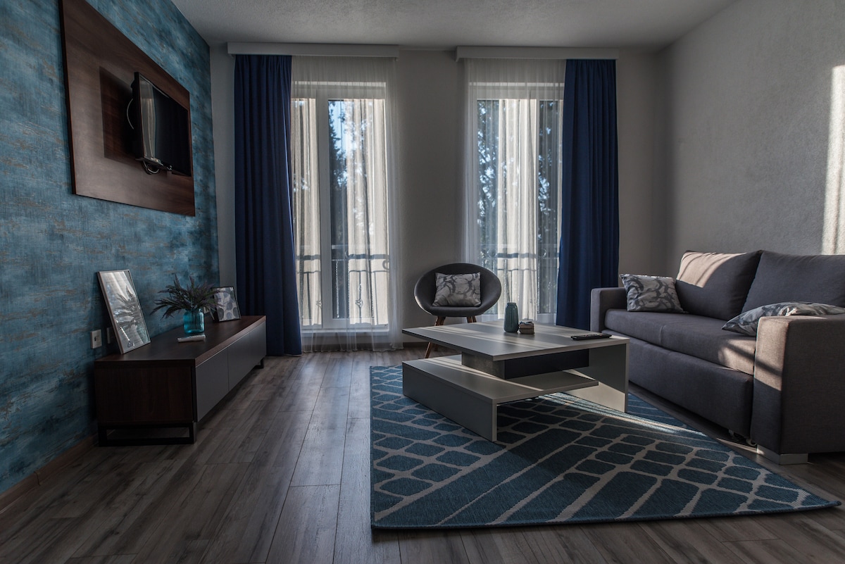 A modern living room is highlighted by a warm color palette, featuring a cozy sofa and a stylish chair. Large windows with sheer curtains allow natural light to fill the space. A geometric rug anchors the coffee table, creating a welcoming environment.
