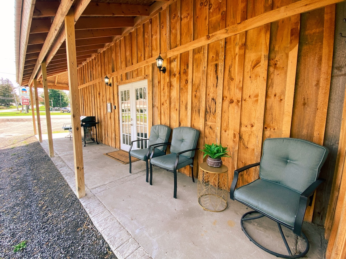 An outdoor seating area features two cushioned chairs beside a small table with a potted plant. The porch is bordered by rustic wooden panels and is well-lit by wall-mounted sconces, providing a welcoming entrance to the space.