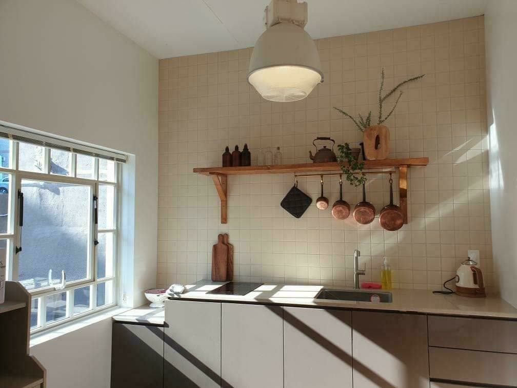 A well-organized kitchen features a mix of natural wood and modern cabinetry. Wall shelves hold various pots and decorative items. A large window provides ample natural light, casting shadows across the countertop, which is equipped with essential appliances and utensils.
