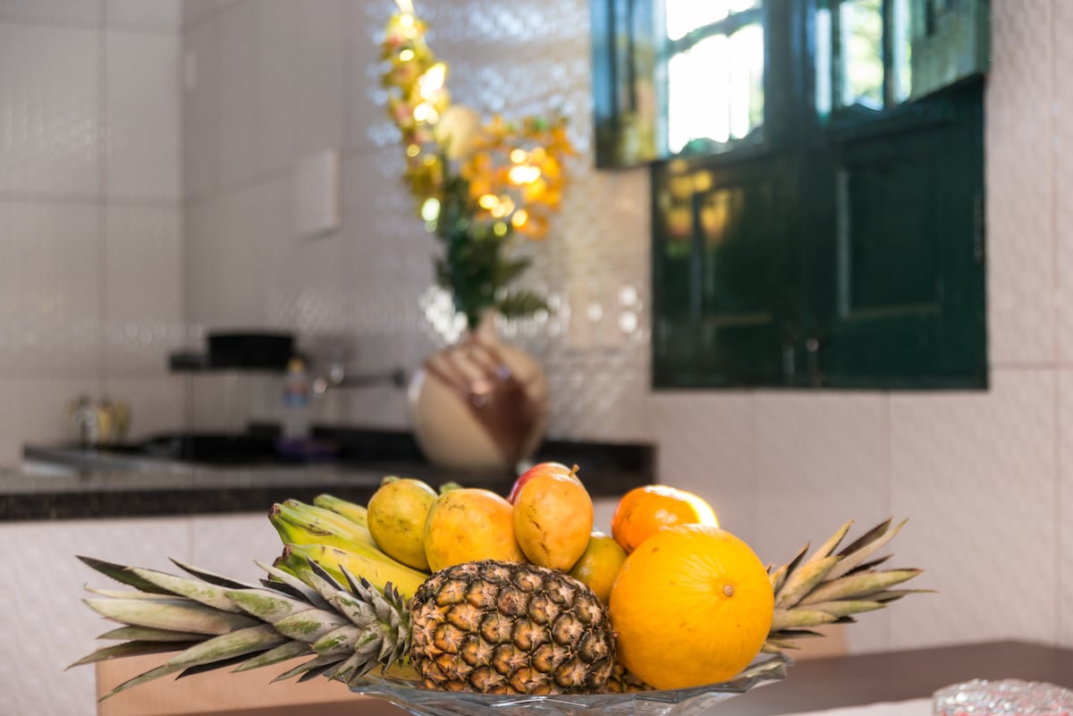 A glass fruit bowl occupies the foreground, filled with a variety of colorful fruits including a pineapple, oranges, and bananas. The kitchen area is visible in the background, featuring tiled walls and a window framed by green shutters, allowing natural light to brighten the space.
