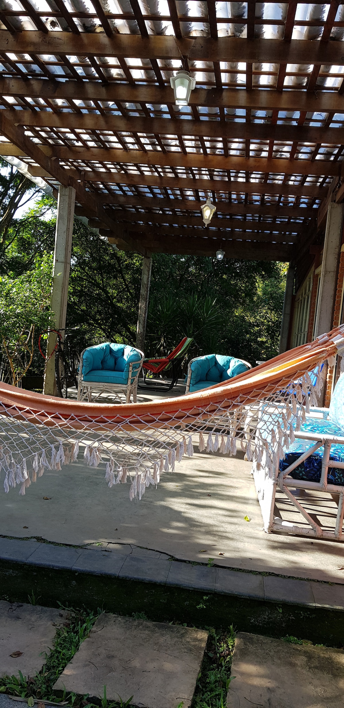 A shaded outdoor patio area features a colorful hammock suspended between posts, with two turquoise armchairs positioned nearby. The surrounding greenery adds to the tranquil environment, while a stone path leads towards the seating arrangement.