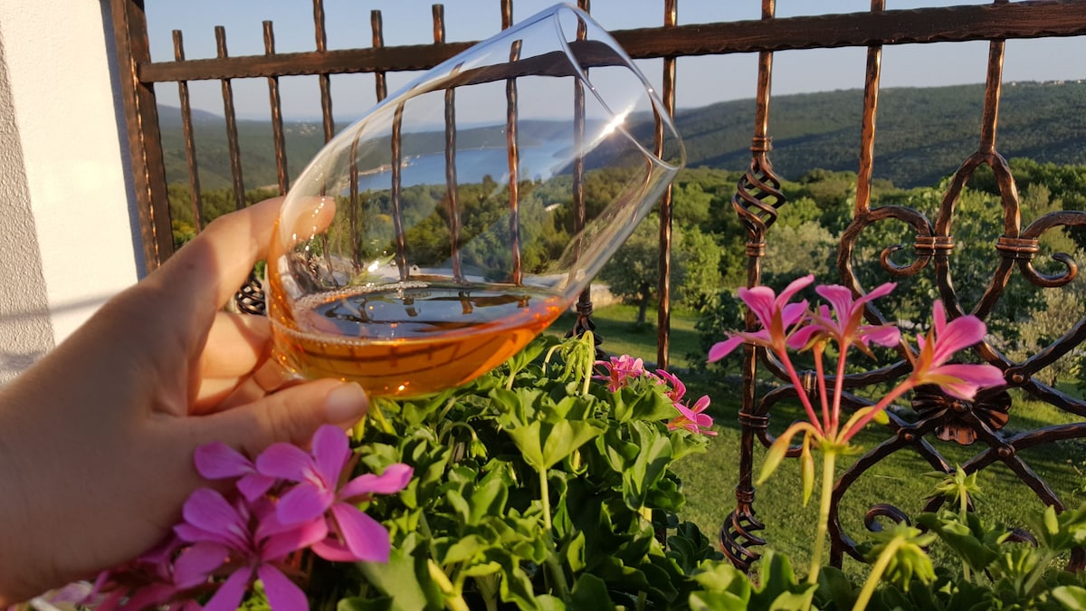 A gently held glass reveals amber liquid against a backdrop of vibrant pink flowers. The view extends to rolling hills and the Rasa Bay, framed by an ornate iron balcony railing. Lush greenery fills the landscape under a clear sky.