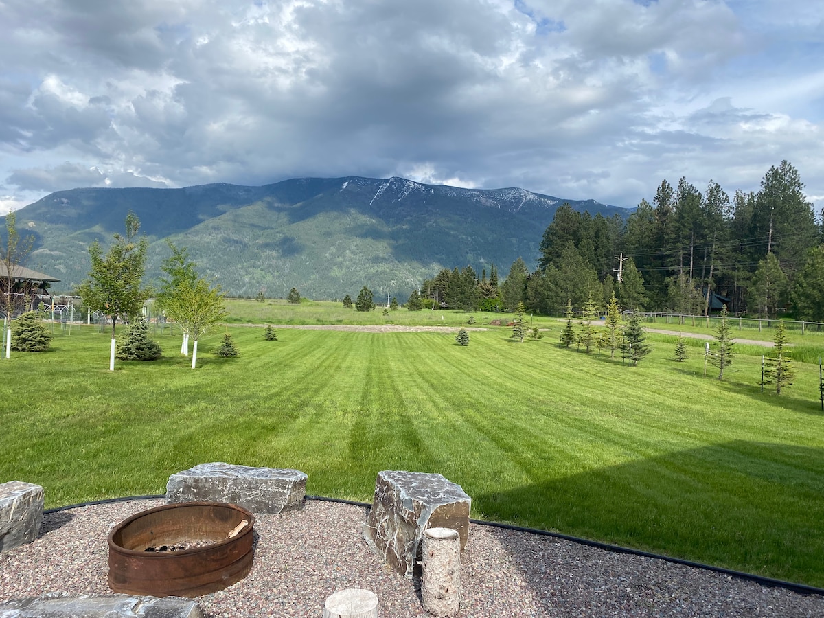 An open lawn area is featured, bordered by neatly trimmed grass and a stone fire pit in the foreground. Majestic mountains rise in the background, under a sky filled with soft clouds, providing a serene outdoor setting.