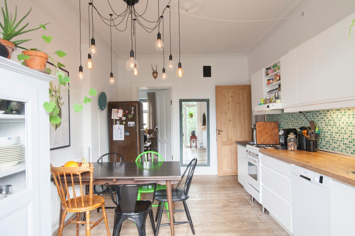 A modern kitchen space features a wooden table surrounded by four chairs, complemented by hanging light bulbs. A tiled backsplash in green hues adds a pop of color. White cabinetry and a wooden countertop provide ample storage and workspace, while a reflection of the dining area is visible in a nearby mirror.
