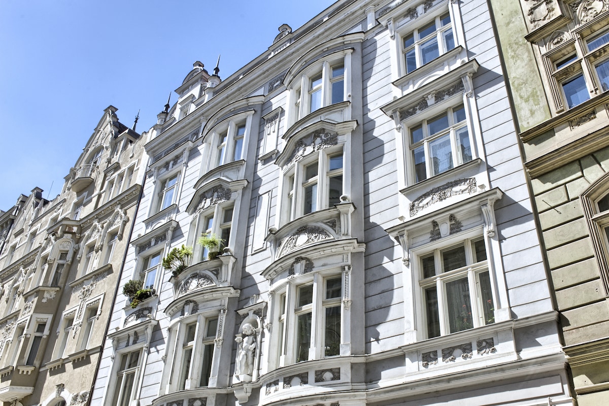 An elegant apartment building is prominently displayed, featuring ornate architectural details on its façade. Multiple windows are visible, accentuated by decorative elements and planters. The clear blue sky provides a bright backdrop, highlighting the building's classic design.