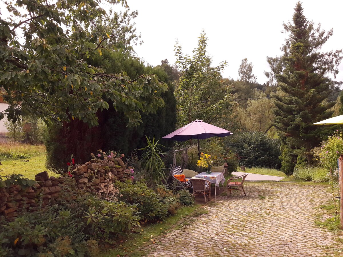 A serene outdoor space is presented with cobblestone pathways leading through greenery. A round table, set for dining, is shaded by a large purple umbrella. Surrounding flora includes flowering plants and shrubbery, creating an inviting atmosphere for relaxation.