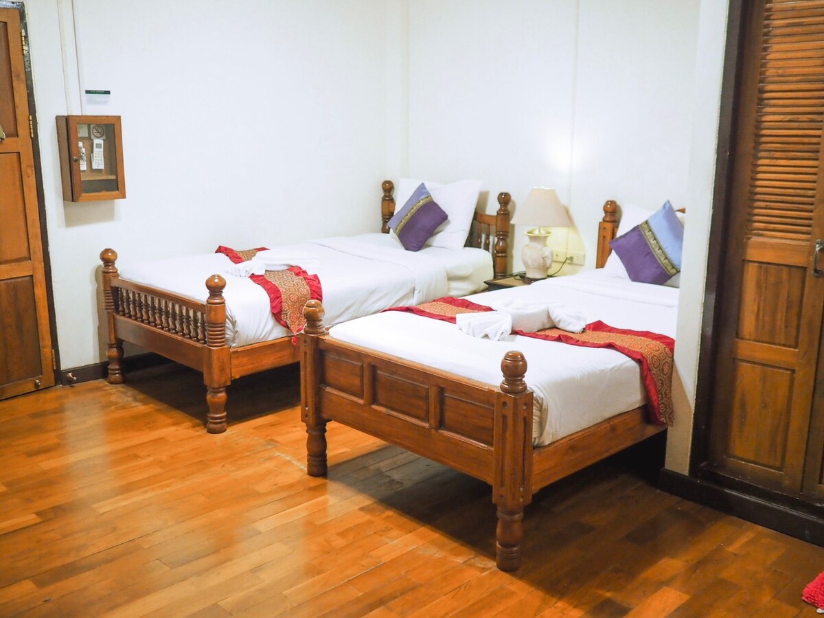 The room features two wooden twin beds dressed in white linens, accented with decorative pillows. A bedside lamp is positioned between the beds. Warm wooden flooring complements the neutral walls, enhancing the spacious feel of the area.