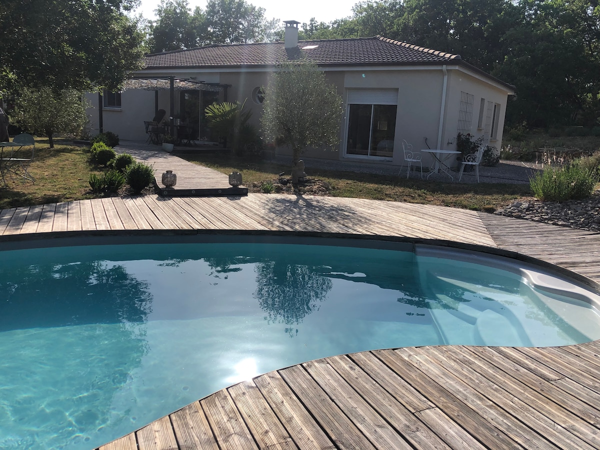 The image captures a serene pool area with curving wooden decking, leading to a light-colored house framed by greenery. Sunlight reflects off the clear water, enhancing the tranquil outdoor setting, while a seating area is visible near the house.